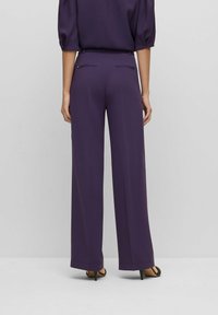 Kalhoty s širokými nohavicemi z tmavě fialového materiálu, s hladkým povrchem, zadními kapsami se zipy a přizpůsobeným střihem, doplněné černými podpatky.