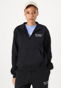 Svart zip-up hettegenser med frontlomme, med "New Balance Athletic Club" logo i hvitt. Brukt over en lys blå skjorte.