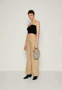 Schwarzes trägerloses Oberteil, beigefarbene weit geschnittene Hose und schwarze Knöchelstiefel. Hält eine kleine, strukturierte, kariert Handtasche. Minimalistisches Design.
