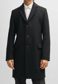Cappotto lungo in lana nera con design raffinato, revers a disegno, sei bottoni e tasche laterali. Tessuto liscio e lunghezza fino al ginocchio.