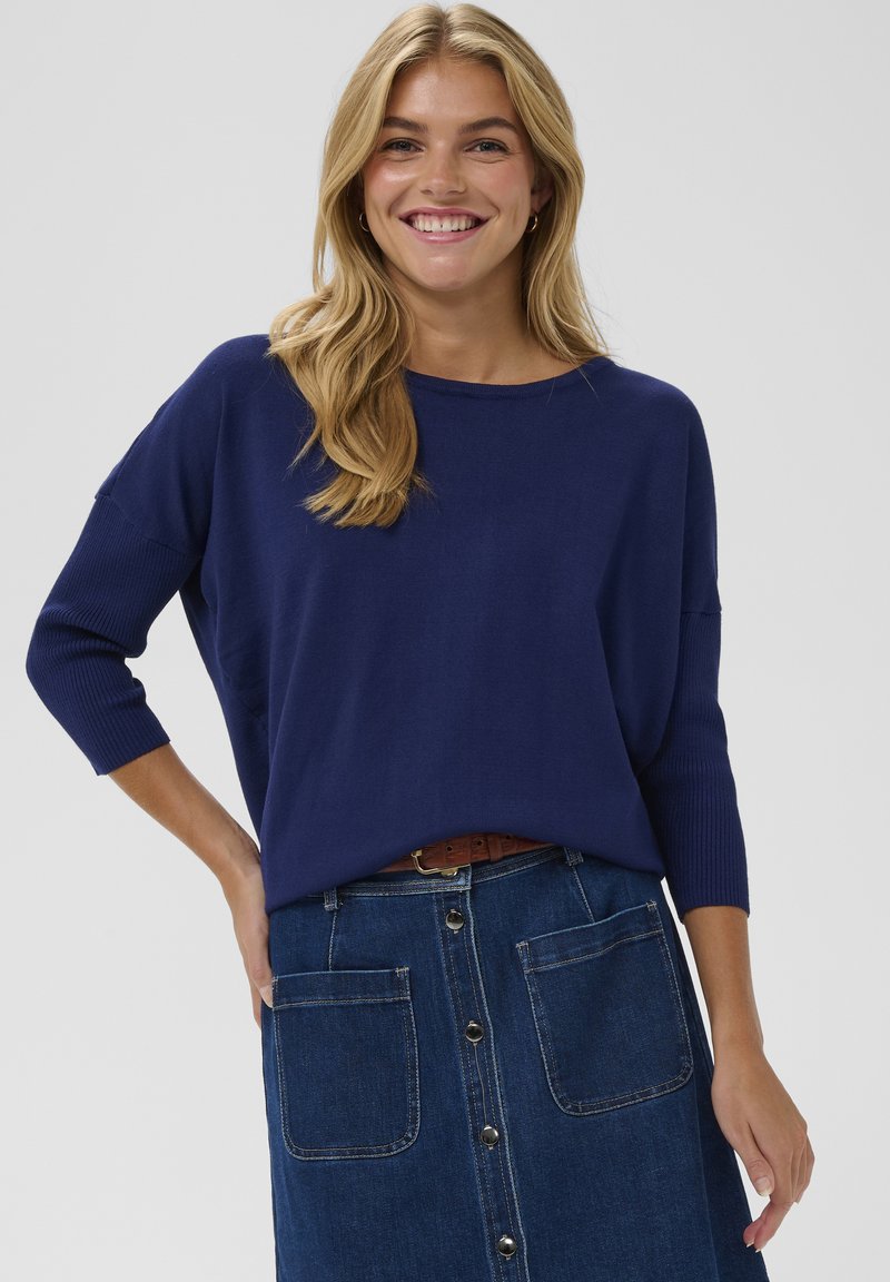 Pull surdimensionné bleu marine avec manches trois-quarts, associé à une jupe en denim dotée de poches avant et d'une fermeture à boutons au centre.