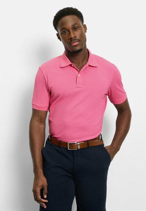 Mann trägt ein leuchtend pinkes Poloshirt mit kurzen Ärmeln, dunkelblaue Hose und einen braunen Gürtel, steht mit einer Hand in der Hosentasche.