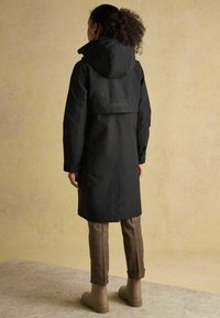 Cabană neagră rezistentă la apă, cu glugă și logo pe spate. Purtată peste pantaloni în carouri maronii și botine bej.