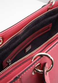 Różowa torba na ramię z teksturowanym wykończeniem, z czarnym wnętrzem, kieszenią zapinaną na zamek i srebrnymi akcentami. Wewnątrz widoczna metka z logo.