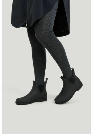 Ben iført mørkegrå leggings og sorte ankelregnstøvler med trækflapper, stående på en lysgrå gulv mod en ensfarvet baggrund.