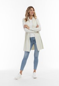 Lys beige lang cardigan med en struktureret front, åben design og ribbede accenter, parret med lyseblå tætsiddende jeans og hvide sneakers.