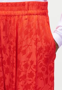 Κόκκινο παντελόνι με ελαστική μέση και διακριτικό λουλουδάτο μοτίβο, φορεμένο με το χέρι στην δεξιά τσέπη και ανοιχτό ροζ μακρυμάνικη μπλούζα.