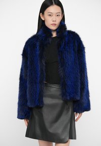 Γυναίκα που φοράει ένα μαύρο τοπ, μαύρη δερμάτινη φούστα και μπλε-μαύρο ψεύτικο γούνινο μπουφάν, στέκεται μπροστά από μονόχρωμο λευκό φόντο.