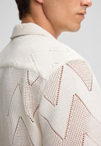 Witte gebreide shirt met een getextureerd zigzagpatroon, notch-kraag en een ademende open weefsel. Zijaanzicht toont schouderdetail.