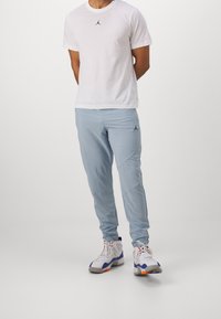 Biała koszulka z czarnym logo, jasnoniebieskie spodnie sportowe z bocznymi szwami oraz białe sneakersy z kolorowymi akcentami. Minimalistyczny design odzieży sportowej.