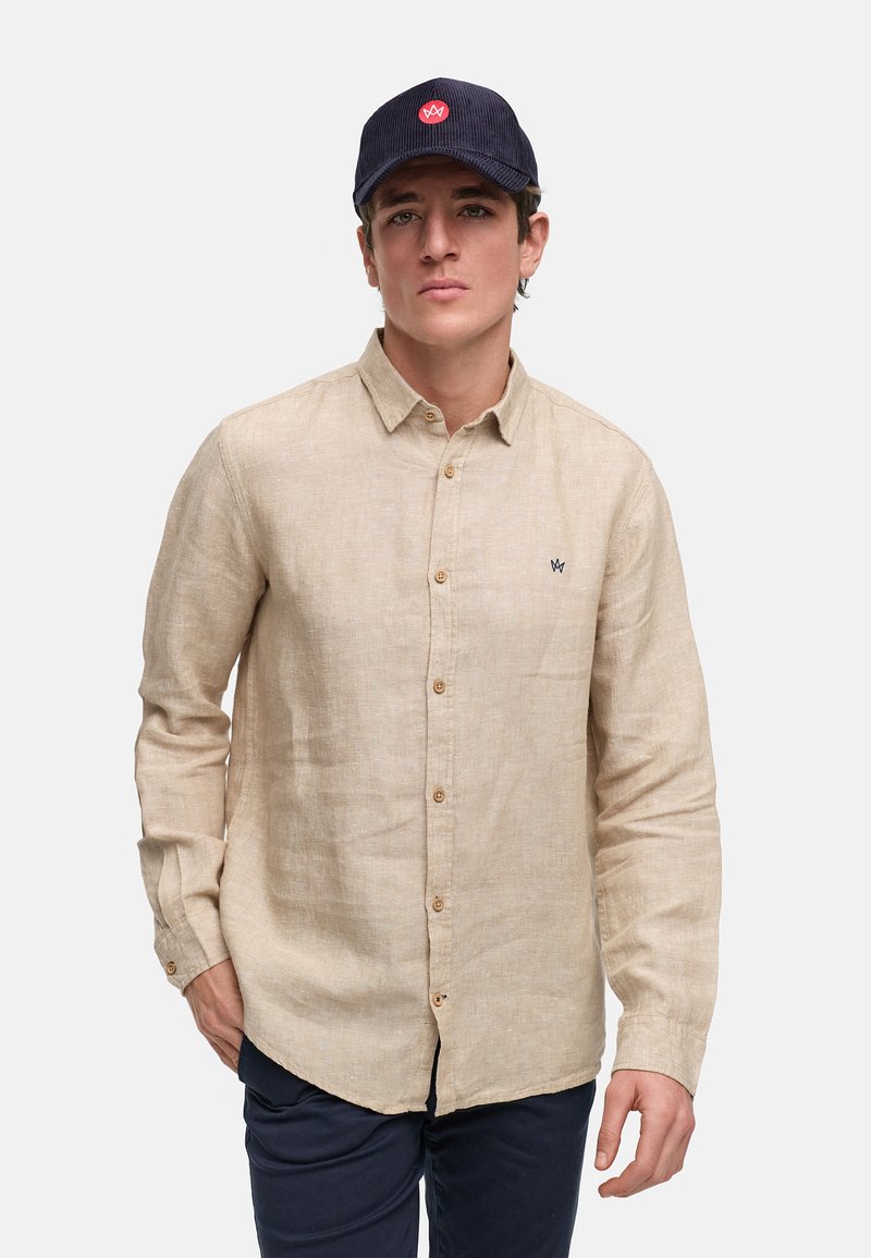 Camisa de lino beige abotonada de manga larga, con un pequeño logo bordado y botones marrones; combinada con una gorra azul marino.