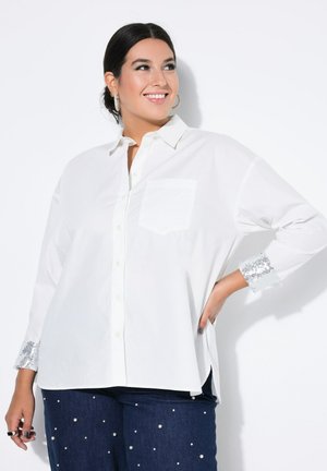 Chemise blanche à manches longues et boutons, avec une poche avant et des accents en sequins argentés sur les poignets. Dotée d'un col et d'une coupe décontractée.