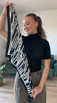 Šal s crno-bijelim uzorkom i geometrijskim prugama, izrađen od glatke tkanine, držan vertikalno. Model nosi crni rebrasti top i bež hlače.