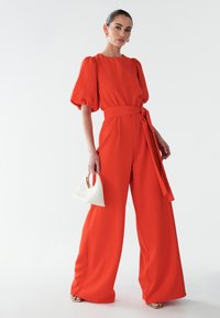 Punane lai jumpsuit puhvärvide ja sidumisega vööl, valmistatud siledast kangast. Kombineeritud väikese valge käekotiga, millel on kullatud käepide.