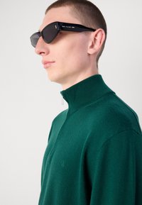 Pullover tricotato verde, strutturato, con colletto alto e zip. Il modello indossa occhiali da sole neri angolari con un discreto marchio sul braccio.