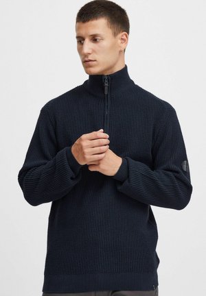 Dunkelblauer Strickpullover mit hohem Kragen und Halbreißverschluss, ausgestattet mit strukturiertem Stoff und kontrastierenden Rippbündchen sowie einem Rippbund am Saum.