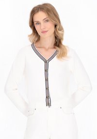 Vrouw met golvend blond haar, gekleed in een witte cardigan met gestreepte afwerking en knopen, in combinatie met witte broek, tegen een lichte achtergrond.
