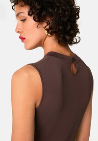 Bruine mouwloze top met ribbeltextuur, hoge halslijn en een keyhole-detail op de rug, bevestigd met een knoop.
