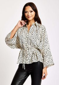 Cream blouse with black speckled pattern, button-up collar, cinched waist, and wide sleeves. Paired with black faux leather pants.