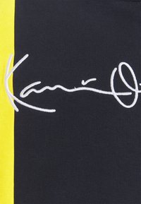 Tejido negro con una franja amarilla vertical; presenta un diseño de firma bordado en blanco. Textura suave, diseño casual.