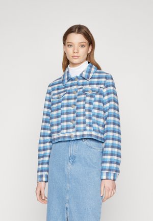 Geknipte jas in een blauw-wit geruite patroon, met een knoopsluiting aan de voorkant, twee borstzakken en een stevige kraag. Onder dit alles een denimrok.