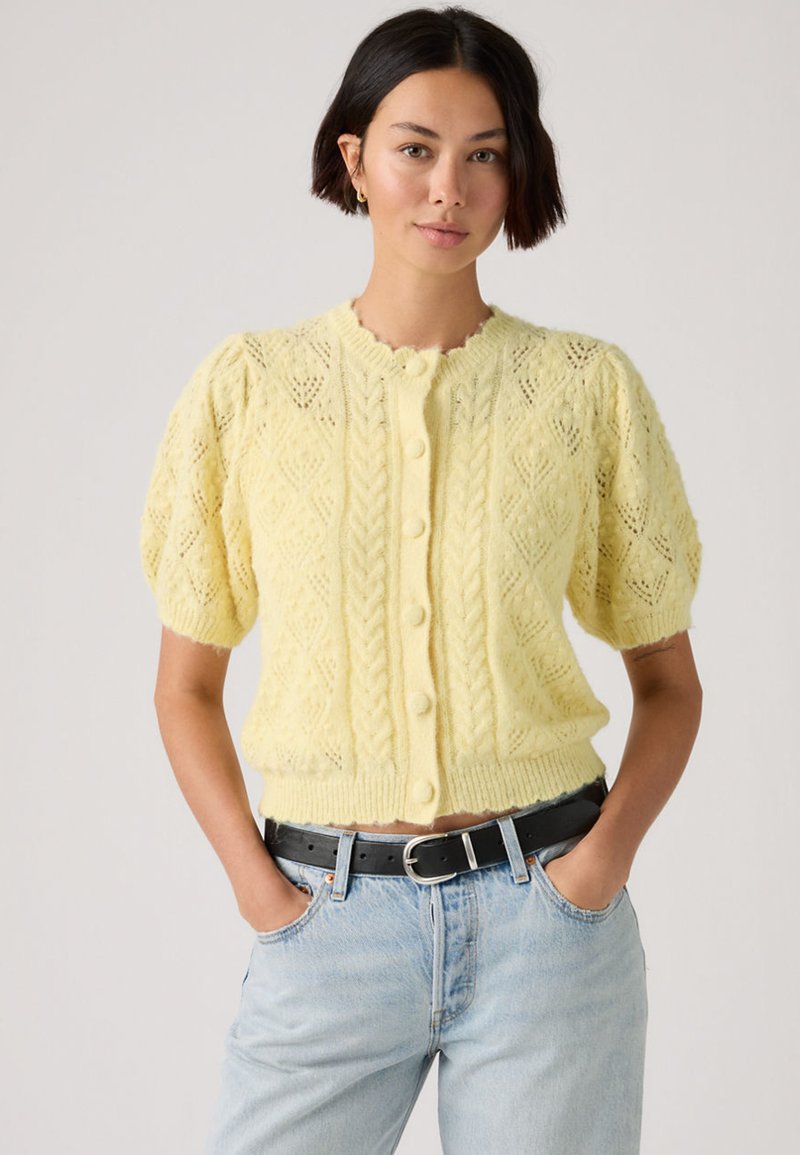Γυναίκα που φορά ένα ανοιχτό κίτρινο, κουμπωτό, πλεκτό ζακέτα με κοντά μανίκια και ανοιχτό μπλε τζιν με μαύρη ζώνη, με τα χέρια στις τσέπες.