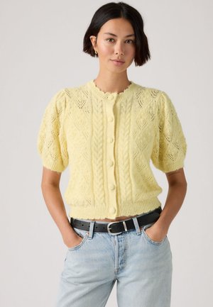 Kvinde iført en lysegul, knappet, strikket kortærmet cardigan og lyseblå jeans med sort bælte, hænderne i lommerne.