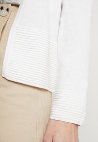 Cardigan bianco lavorato a maglia con polsini e orlo a coste, texture morbida, abbinato a un tessuto beige testurizzato sotto.