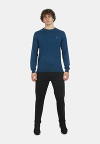 Maglione azzurro a maniche lunghe realizzato in un tessuto morbido, con scollatura a girocollo, polsini a coste e un piccolo logo sul petto. Abbinato a pantaloni neri.