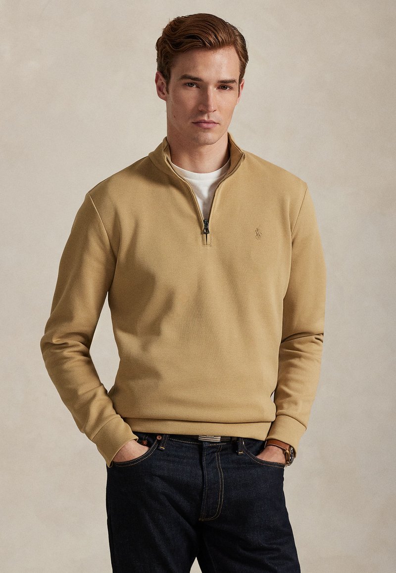 Νεαρός άνδρας με ανοιχτό καστανά μ αλλιά, ντυμένος με μπεζ πουλόβερ με φερμουάρ στο λαιμό πάνω από λευκό πουκάμισο και σκούρο μπλε τζιν, με τα χέρια στις τσέπες.