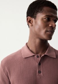 Korte mouw, geribde poloshirt in mauve met een kraag en drie bruine knopen. Gladde textuur en een aangesloten ontwerp, getoond op de nek en schouder van een model.