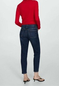 Tmavě modré džíny z denimu s přiléhavým střihem, zadními kapsami a zkrácenou délkou, kombinované s červeným topem s dlouhým rukávem a černými sandály na podpatku.