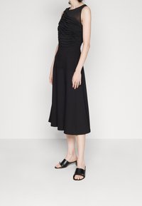 Femme portant une robe midi noire sans manches avec un corsage froncé et un décolleté transparent, associée à des mules noires à bout ouvert sur un fond uni.