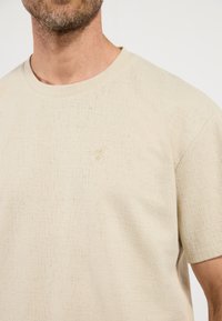 Beige tričko s krátkým rukávem vyrobené z texturované látky, s malým zlatým ptáčkem na levé hrudi. Kulatý výstřih, volný střih.