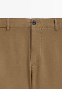 Brązowe spodnie w talii z szlufkami na pasek i czarnym zapięciem na guzik na jednolitym białym tle.