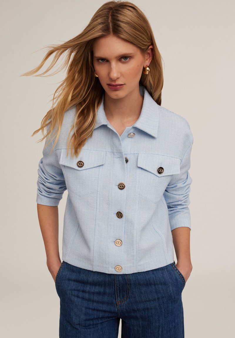 Giacca azzurro chiaro realizzata in tessuto texture, con colletto, due tasche sul petto e chiusura con bottoni dorati. Abbinata a jeans blu.