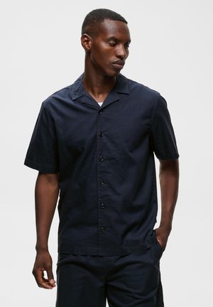 Homme portant une chemise bleu marine foncé à manches courtes avec col et pantalon assorti, debout avec une main dans la poche.