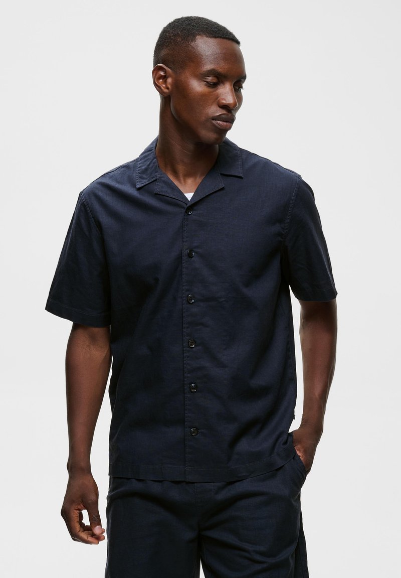 Homme portant une chemise bleu marine foncé à manches courtes avec col et pantalon assorti, debout avec une main dans la poche.