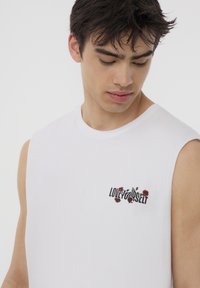 T-shirt bianco senza maniche in cotone con una grafica floreale e la scritta "AMA TE STESSO" in nero e rosso, texture morbida, scollo rotondo.