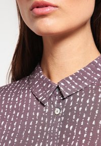 Lila skjorta med krage och ett vitt abstrakt mönster, med knapplåsning och en strukturerad tygkvalitet, sedd från halsringningen och upp.