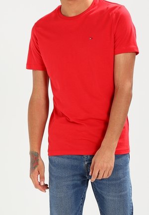 Mand iført en almindelig rød T-shirt med korte ærmer og blå jeans, stående med sin venstre hånd delvist i lommen.