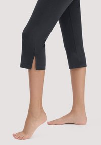 Barfuß, ein Fuß flach auf dem Boden, der andere auf den Zehen, trägt schwarze verkürzte Hosen mit kleinen Seitenschlitzen oberhalb der Knöchel.