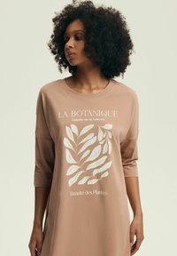 Kobieta z kręconymi włosami, ubrana w jasno brązową sukienkę z białymi roślinnymi wzorami i francuskim tekstem z przodu, patrząca na bok.