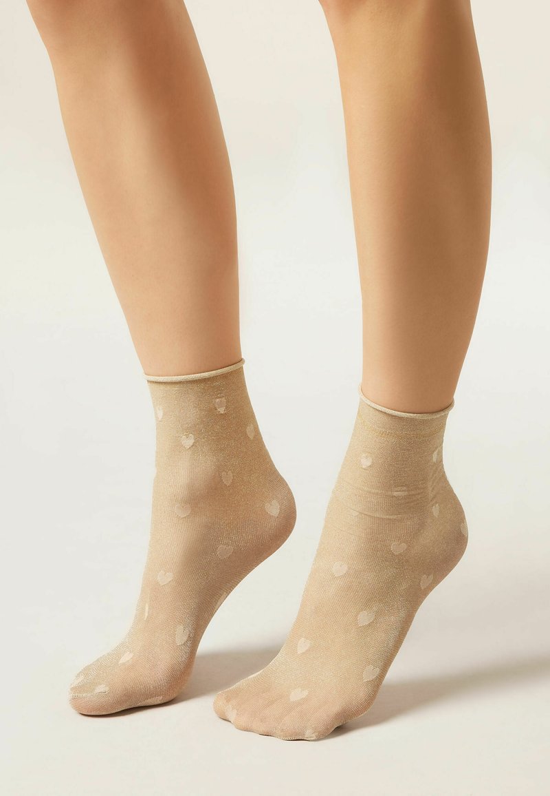 Beine mit durchsichtigen beigefarbenen Knöchelsocken mit kleinen Herzmustern, auf hellem neutralem Hintergrund.