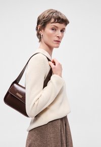 Καφέ δερμάτινη τσάντα ώμου με ορθογώνιο σχήμα, ανάγλυφο σχέδιο και λεπτομέρεια από χρυσό τόνο. Φοριέται με ένα κρεμ πουλόβερ και καφέ φούστα.