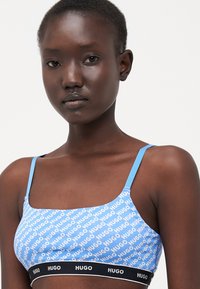 Light blue bralette with repeating white "HUGO" text pattern, features thin straps and a black underband with "HUGO" branding.