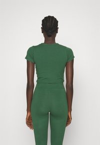 Žena s pletenými vlasy svázanými do drdolu, oblečená v těsném zeleném krátkém rukávu a vysokých zelených legínách, stojící před jednobarevným pozadím.