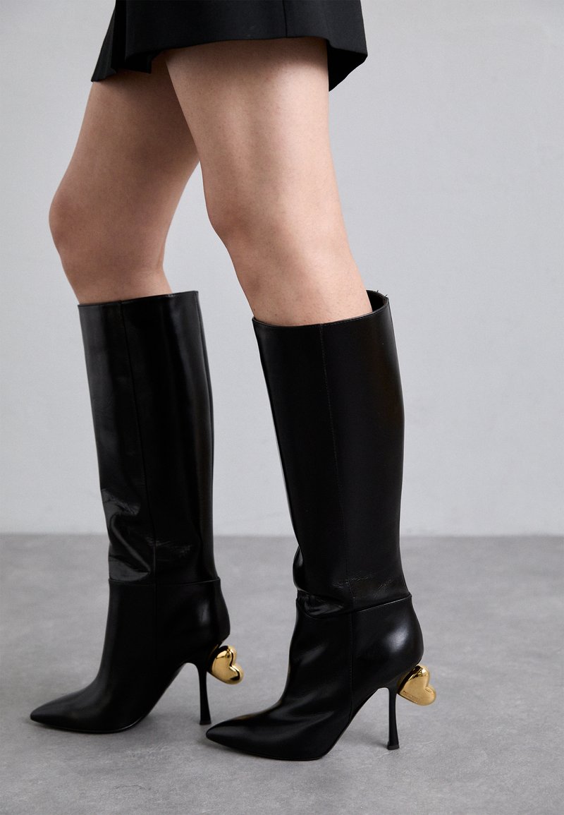 Botine din piele neagră, până la genunchi, cu vârfuri ascuțite și toc stiletto. Prezintă accente aurii în formă de inimă pe toc și o textură netedă, lucioasă.
