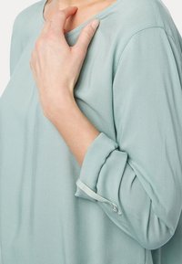 Ljust grön blus med lång ärm och rundad halsringning, mjukt tyg och uppvikta ärmar. En anmärkningsvärd detalj är en knappdetalj på manschetten.