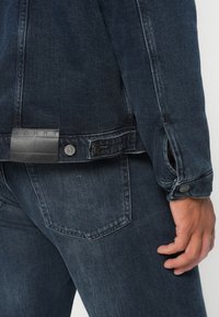 Krupan plan osobe koja nosi tamnoplavu traper jaknu i traperice, prikazujući stražnji džep, rub jakne s metalnim dugmadima i dio desne ruke.